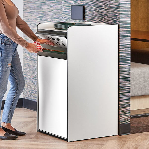 A woman using a white Lancaster Table & Seating waste receptacle with a black "THANK YOU" swing door to throw away paper.