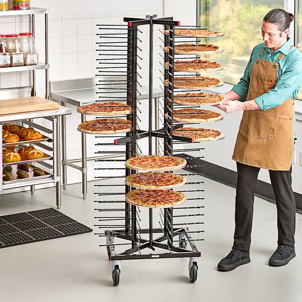 A man in an apron standing next to a JackStack mobile pizza plate rack filled with pizzas.