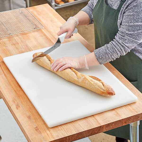 A person cutting a baguette on a white polyethylene cutting board.