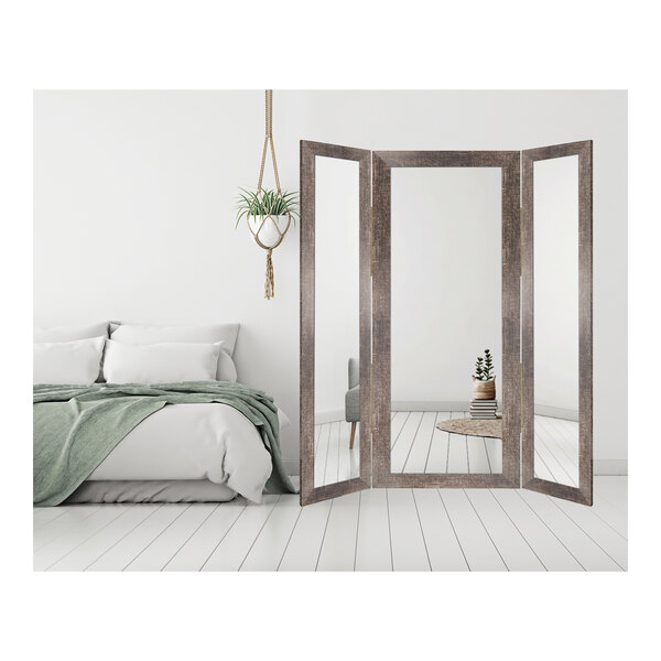 A BrandtWorks trifold dressing mirror with a wooden frame in a room with a bed and a plant.