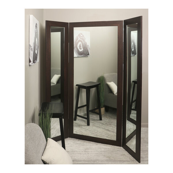 A BrandtWorks trifold dressing mirror in a corner with a stool and pillows.