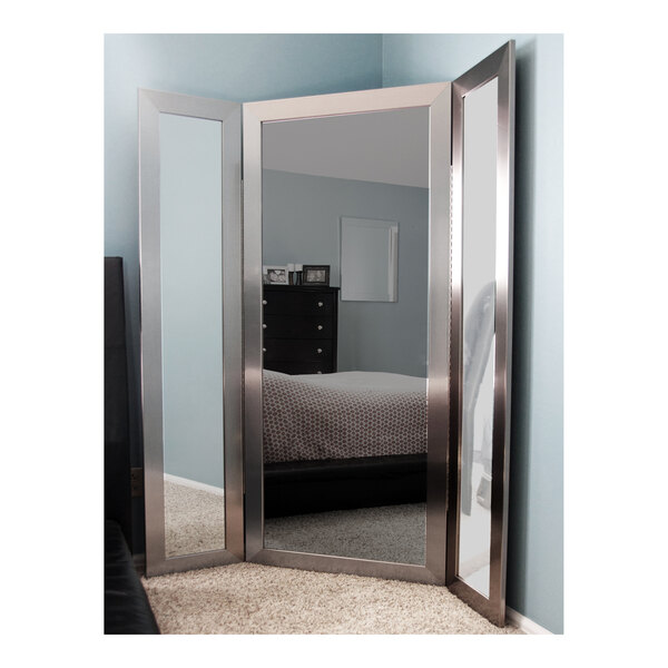 A BrandtWorks silver trifold dressing mirror in a white room above a bed with a red and white blanket.