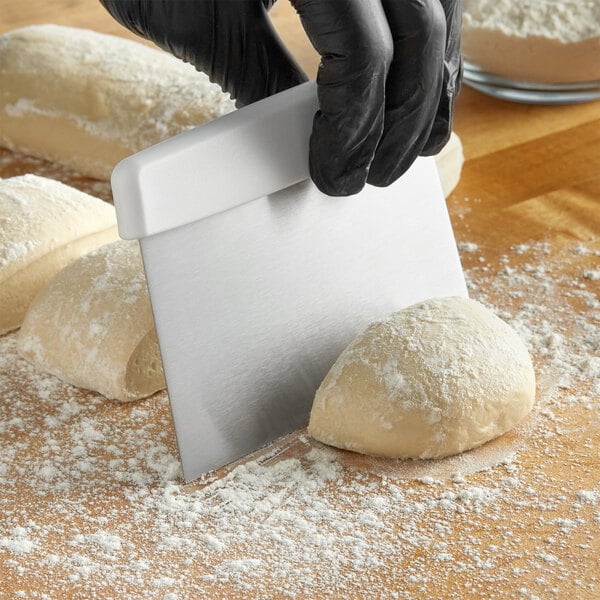 A person in black gloves uses a Choice stainless steel dough cutter with a white handle to cut white dough on a brown table.