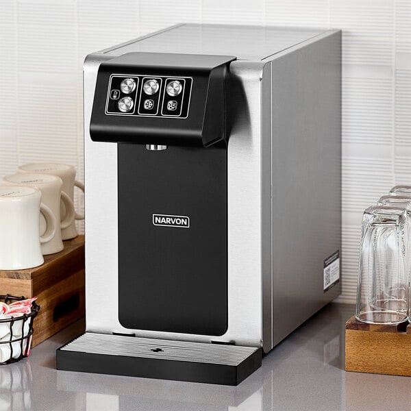 A silver and black Narvon countertop water dispenser next to a clear glass of water.