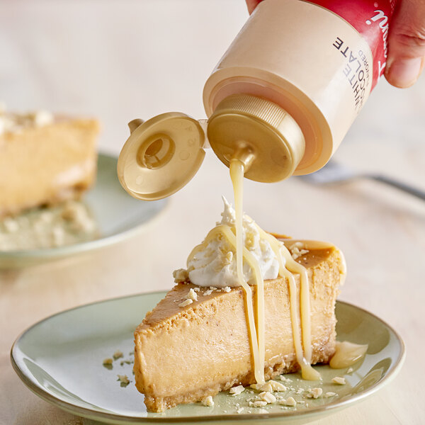 A person pouring white chocolate sauce onto a slice of cheesecake.