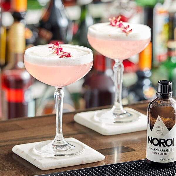 A bottle of NOROI Vegan Cocktail Foamer on a bar counter with two glasses of pink liquid.