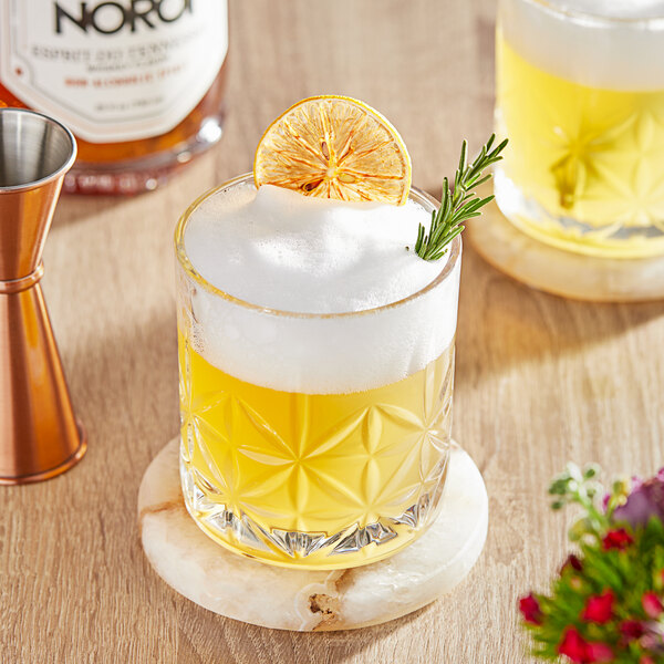 A glass of NOROI non-alcoholic whiskey with a slice of orange and rosemary on a table.