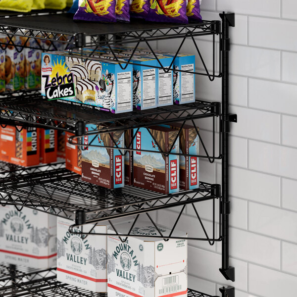 A Regency black wire wall-mount shelf with snacks on it including a box of chocolate bars.