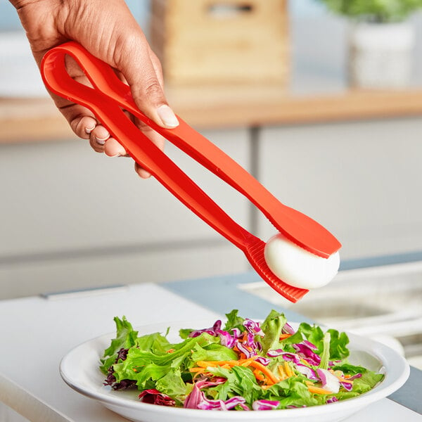 A hand holding a red Choice 9" Polycarbonate Flat Grip Tong over a plate of salad.