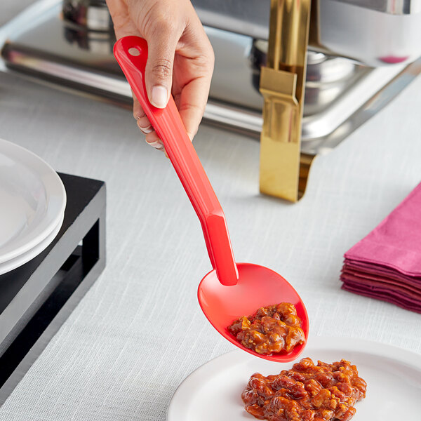 A hand holding a red Choice salad bar spoon over a plate of food.