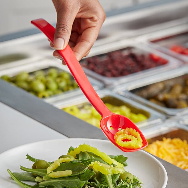 A hand holding a Choice red polycarbonate perforated salad bar spoon over a plate of salad.