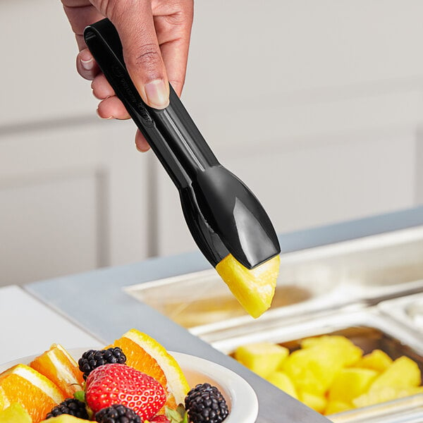 A hand using black polycarbonate salad tongs to serve fruit into a bowl.