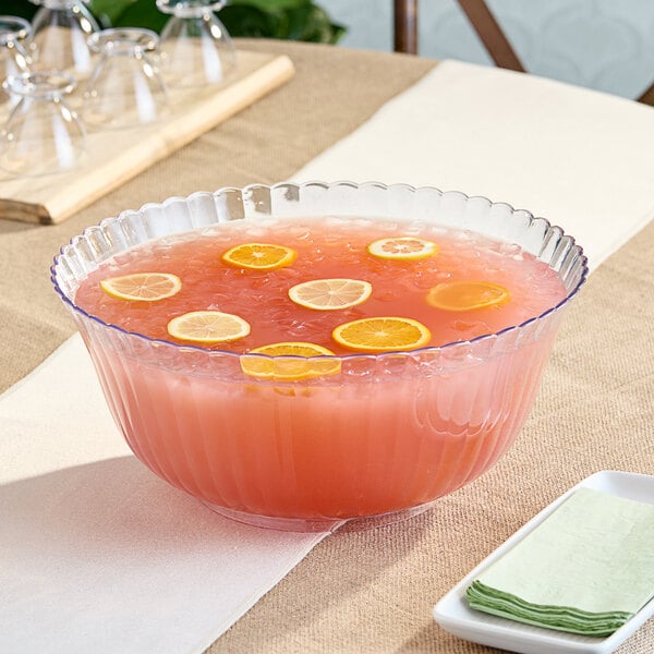 A clear plastic punch bowl of pink liquid with lemons and slices of oranges on a table.