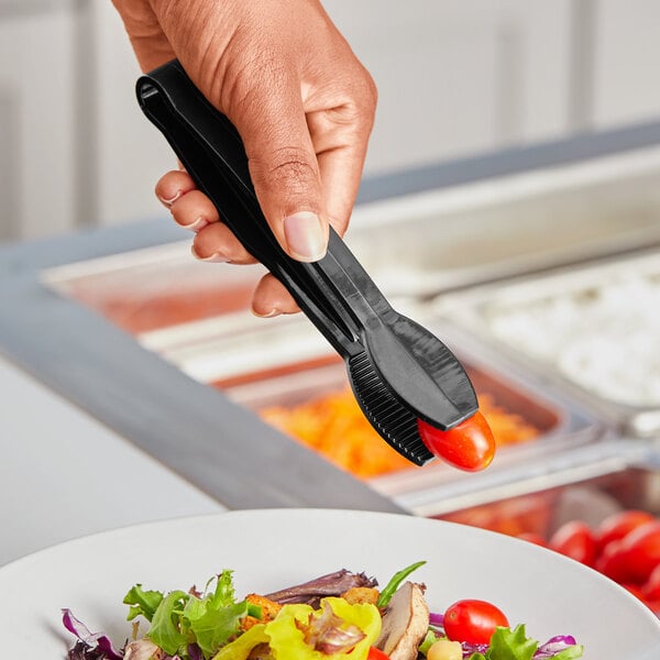 A hand holding a black polycarbonate flat grip tong over a plate of food on a table.