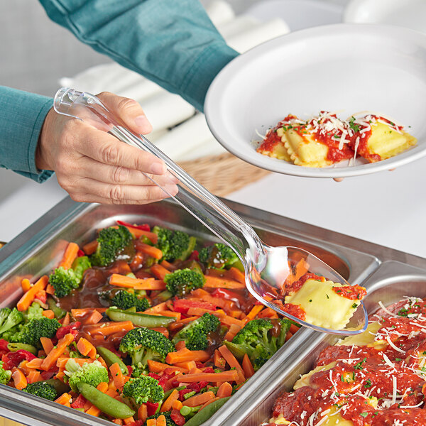 A person using a Visions clear plastic serving spoon to serve vegetables onto a plate.