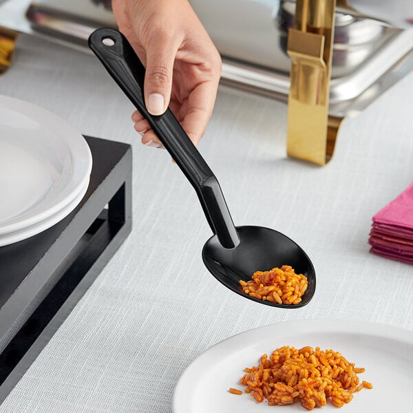 A hand holding a black Choice polycarbonate salad bar spoon over a plate of food.