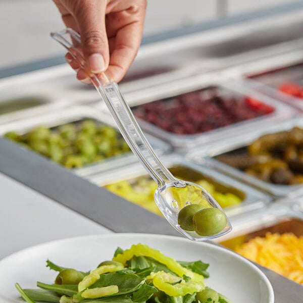 A hand holding a Choice clear polycarbonate perforated salad bar spoon over a plate of salad