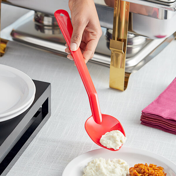 A hand holding a red Choice salad bar spoon over a plate of food.