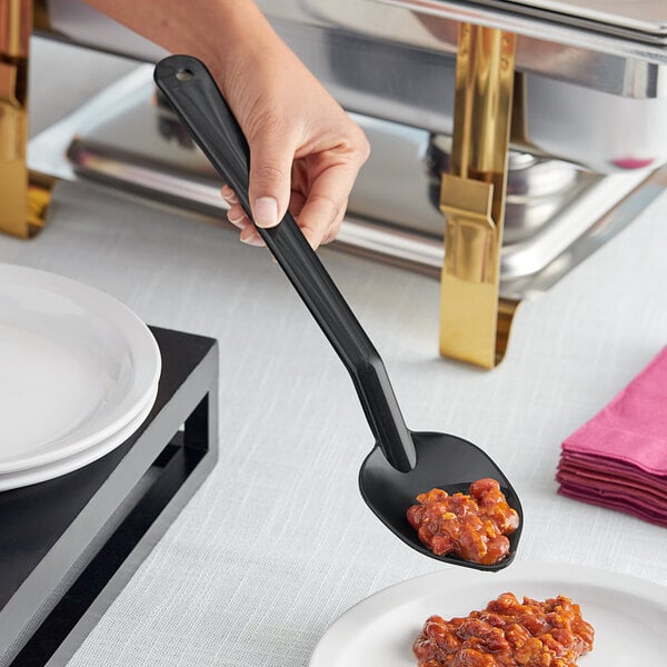 A hand holding a black Choice buffet spoon over a plate of food.