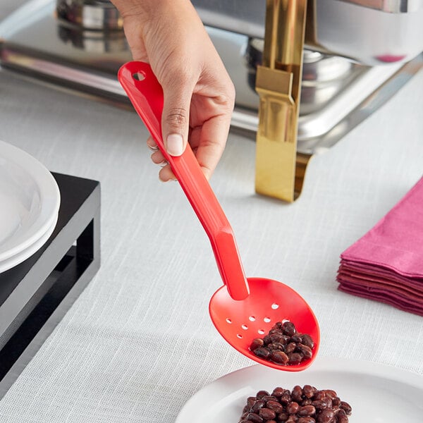 A person holding a red Choice polycarbonate perforated salad bar spoon over a plate of beans.