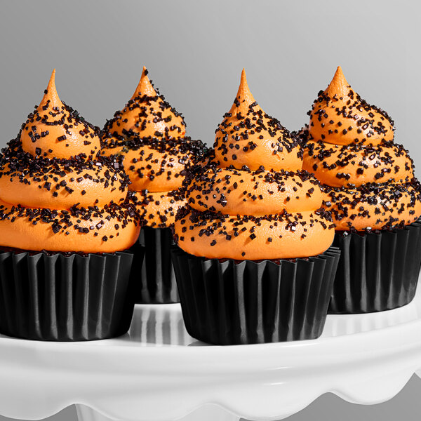 A group of cupcakes decorated with orange frosting and coarse black sanding sugar, displayed on a white stand.