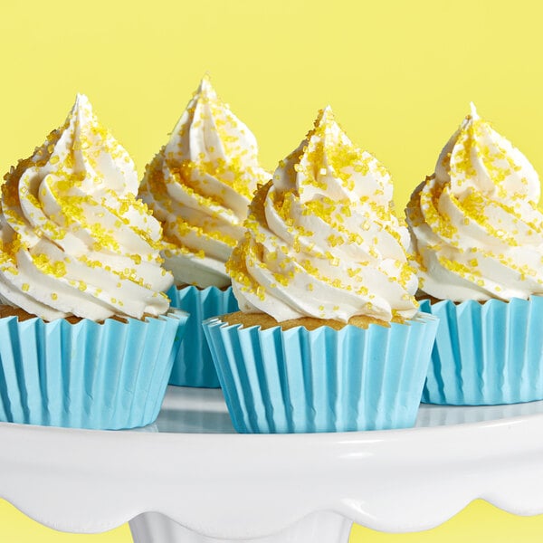 A group of cupcakes with white frosting and yellow coarse sanding sugar in blue wrappers on a white cake stand.