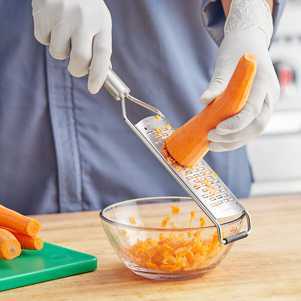 A person in gloves using a Microplane ribbon grater to grate a carrot.