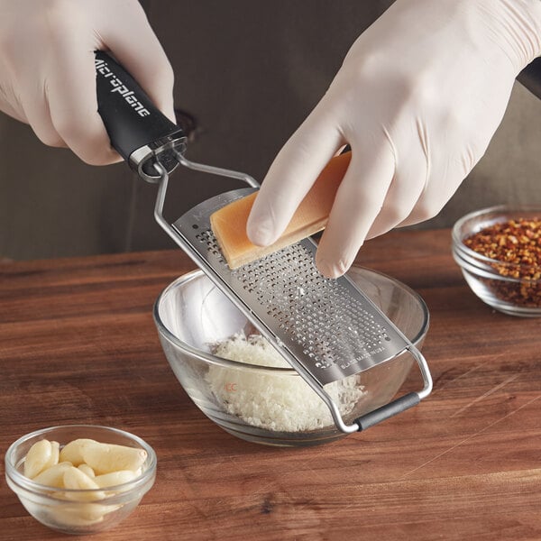 A person in gloves using a Microplane grater to grate cheese in a bowl.