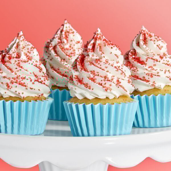 A group of cupcakes with white frosting and red sanding sugar sprinkles in blue wrappers displayed on a white cake stand.