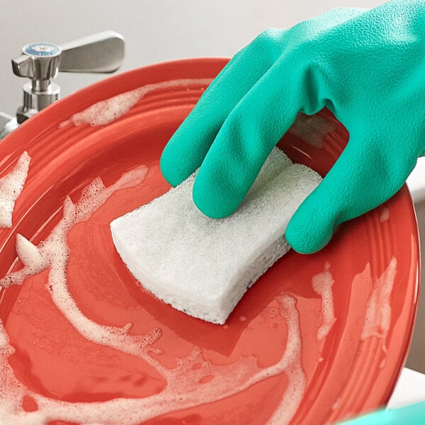 A gloved hand using a 3M Scotch-Brite Greener Clean Non-Scratch Scrub Sponge to clean a red plate.