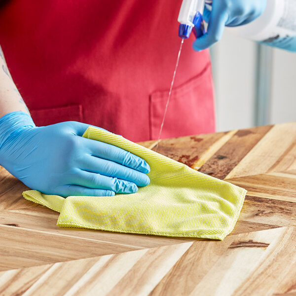 A person wearing blue gloves using a yellow 3M Scotch-Brite microfiber cloth to clean a surface.