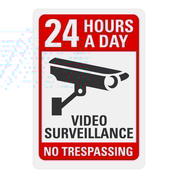 A white rectangular sign with black and red text and a black and white camera symbol. Text reads: "24 Hours A Day / Video Surveillance / No Trespassing"