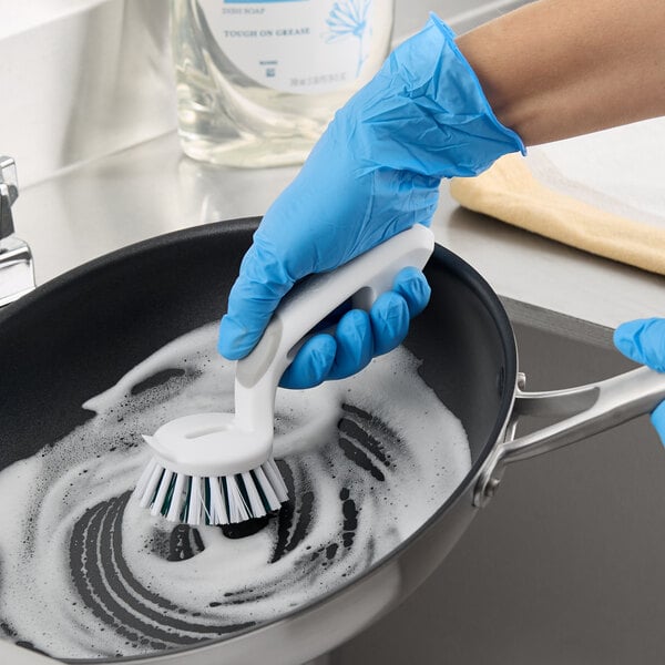 A white and blue scrubber brush with a curved handle being used to clean a soapy frying pan.