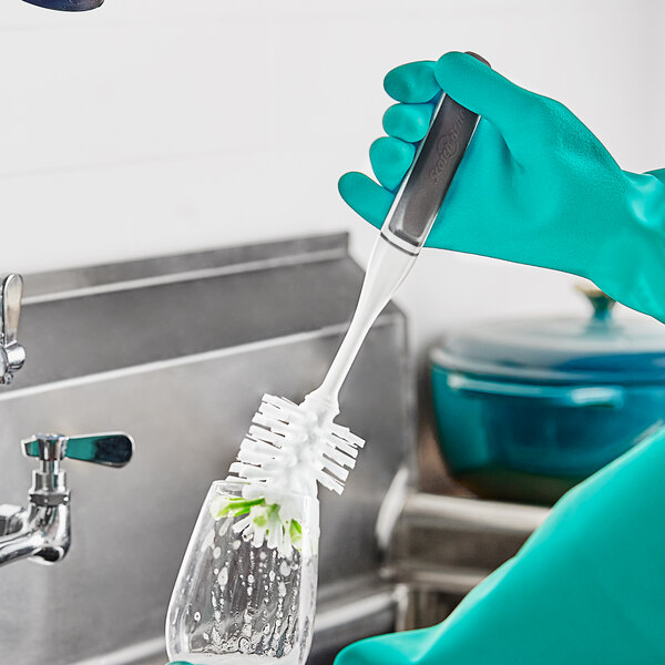 A person in green gloves using a 3M Scotch-Brite Glass Brush to clean a glass over a sink.