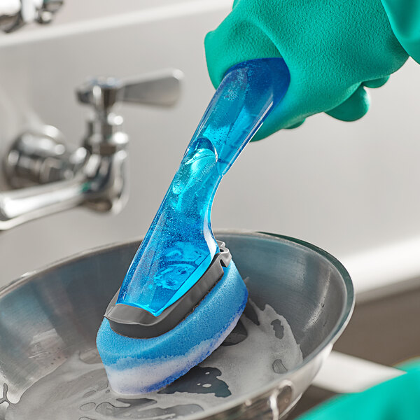 A person washing dishes with a 3M Scotch-Brite blue soap dispensing sponge.