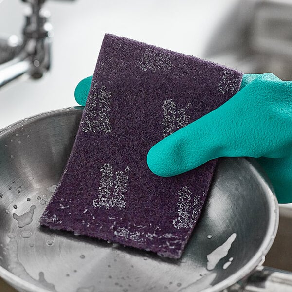 A gloved hand using a 3M Scotch-Brite Extreme Scrub sponge to clean a stainless steel sink.
