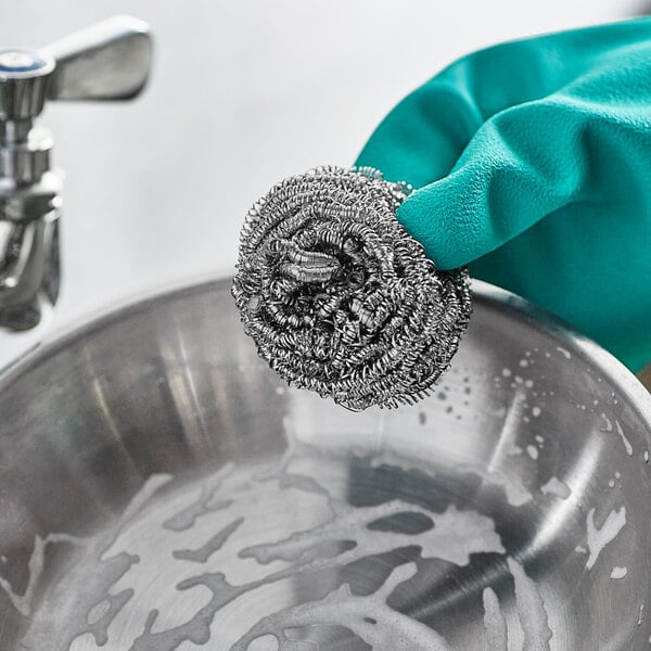 A gloved hand holding a 3M Scotch-Brite stainless steel scrubber over a metal bowl.