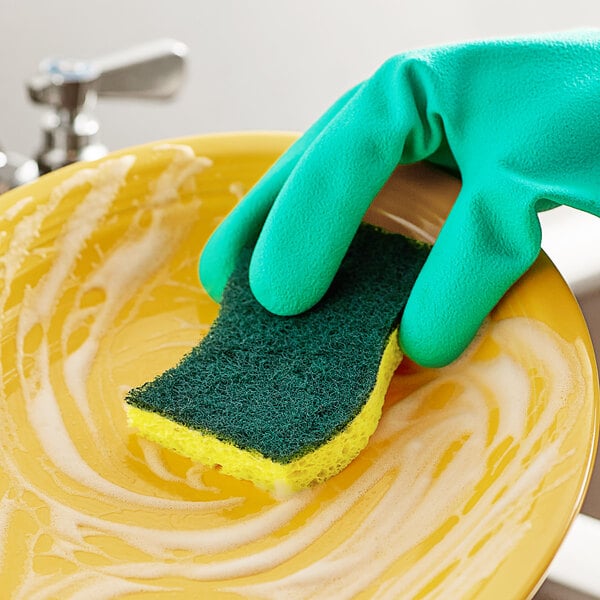 A gloved hand using a 3M Scotch-Brite heavy-duty scrub sponge to clean a yellow plate.