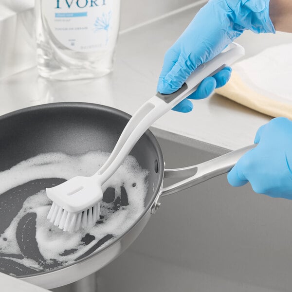 A white 3M Scotch-Brite pot and pan brush being used to clean a soapy frying pan.