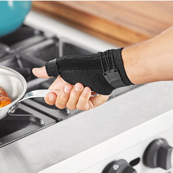 A person wearing a 3M Futuro Deluxe Thumb Stabilizer over a black glove holding a pan.