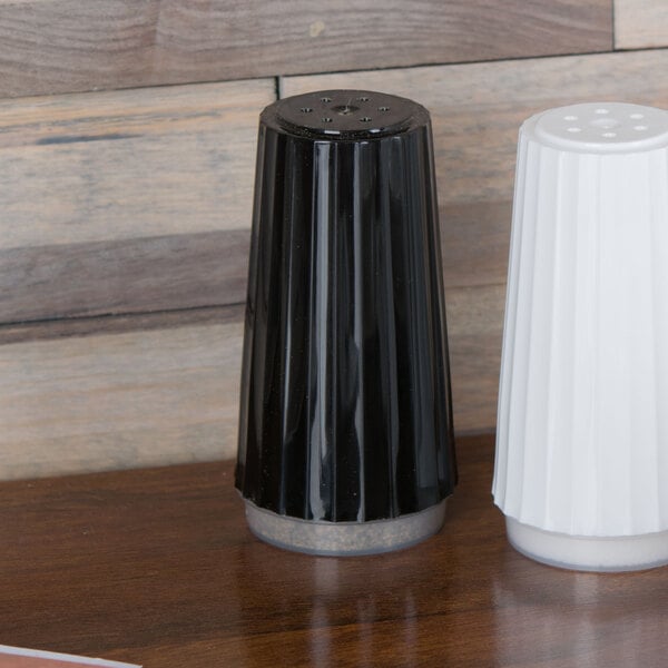 A black container of white pepper on a table with a white column and a black and white pepper shaker.
