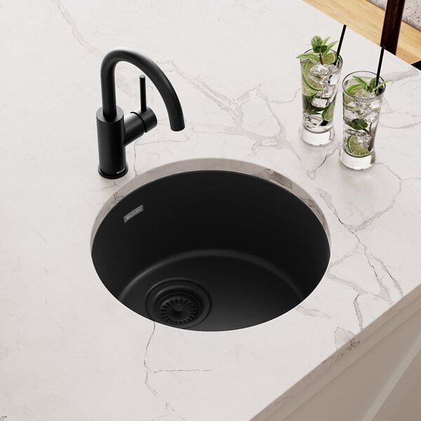 A Zurn Elkay black round Quartz sink with a hole in it on a marble counter with a glass of ice and lime slices.