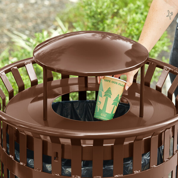 A hand putting a paper cup in a brown Lancaster Table & Seating trash can lid on a outdoor patio table.