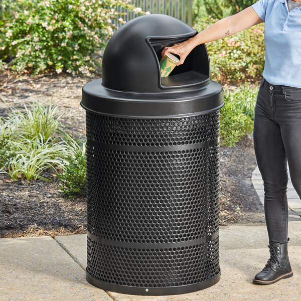 A woman opening a Lancaster Table & Seating outdoor trash can with a plastic dome lid.