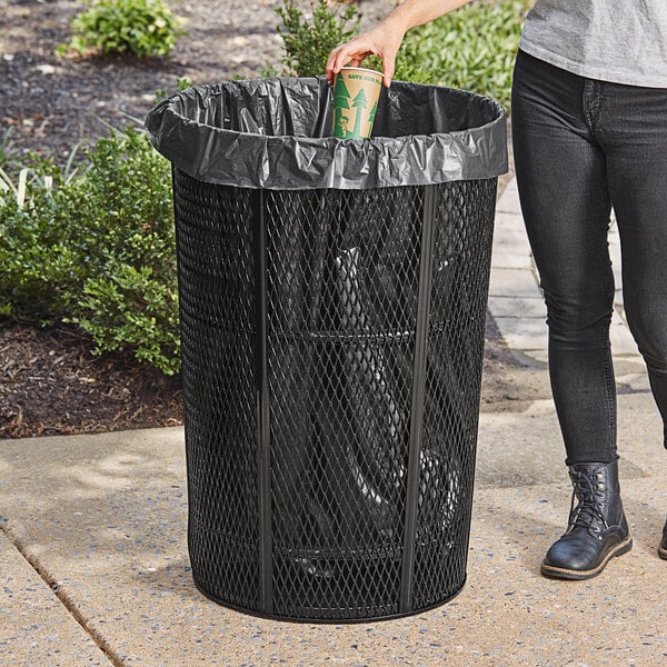 A woman standing next to a Lancaster Table & Seating black steel mesh outdoor trash can and putting a green bottle in it.