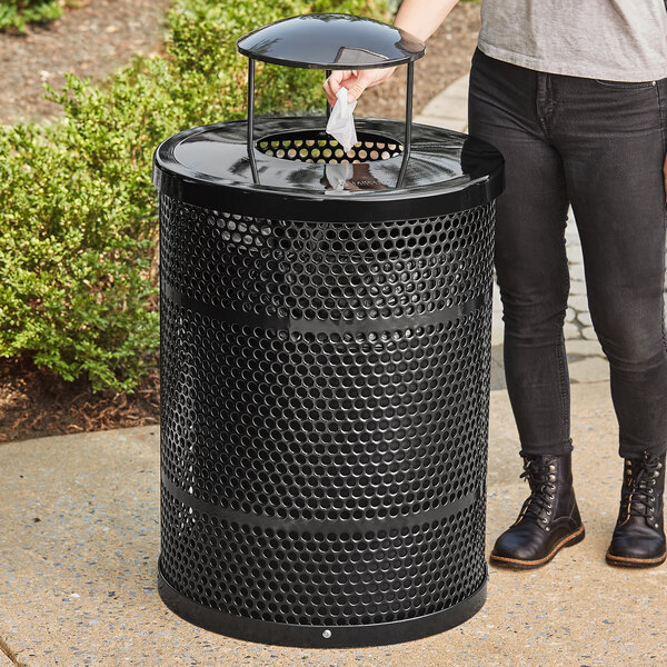 A person standing next to a Lancaster Table & Seating heavy-duty black steel outdoor trash can with a rain bonnet lid.