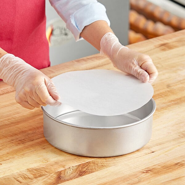 A person wearing gloves places a round white Quilon parchment liner in a metal cake pan.