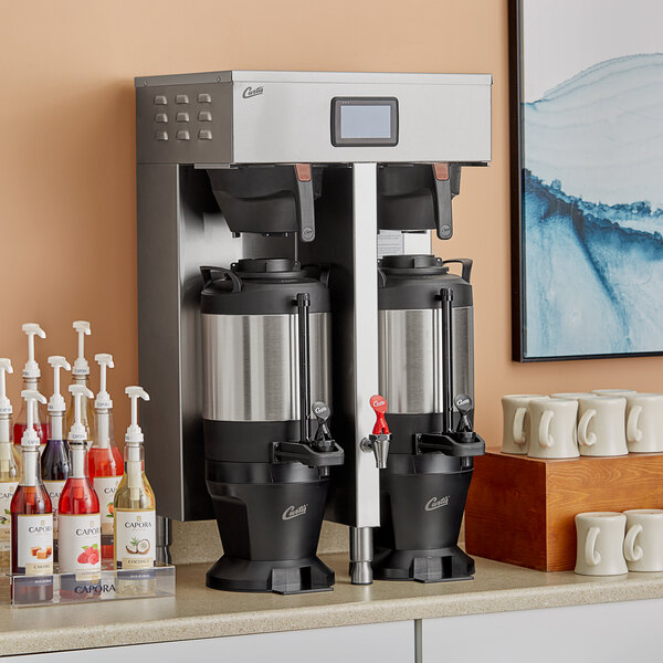 A Curtis stainless steel twin coffee brewer on a counter with white coffee mugs.