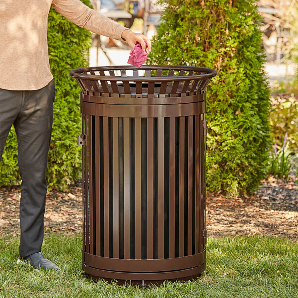 A man standing next to a Lancaster Table & Seating heavy-duty brown steel slatted outdoor trash can with flat lid and door.