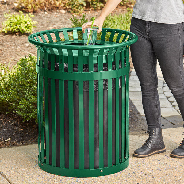 A woman standing next to a green Lancaster Table & Seating outdoor trash can.